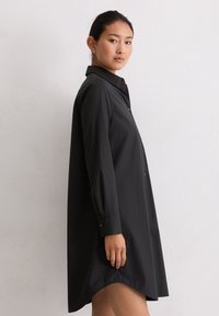 Femme aux cheveux noirs en chignon portant une robe chemise noire à manches longues avec des boutons, se tenant devant un mur blanc texturé.