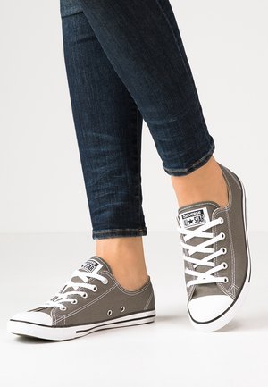 Persona que lleva vaqueros pitillo azul oscuro y zapatillas Converse All Star bajas grises con cordones blancos y suelas de goma.