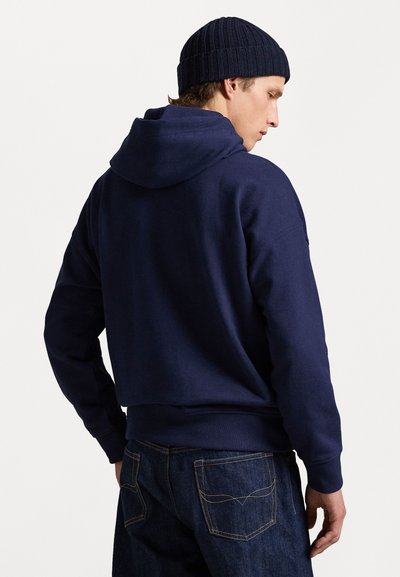 Temno modra kapuca z vrvico, z rebrastimi manžetami in robom; v kombinaciji s temnimi jeansi z vidnimi šivi.