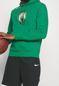 Personne portant un sweat à capuche vert des Boston Celtics et un short noir Nike tenant un basket contre un fond uni.