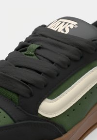Sort og grøn sneakers med en tekstureret læderoverdel, flade snørebånd og en cremefarvet sidelinje. Brun gummisål med ventilationshuller.