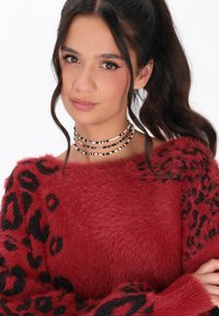 Maglione rosso e morbido con dettagli a stampa leopardata nera sulle maniche. Presenta un ampio scollo e una texture soffice. Collana con molteplici fili di perle visibile.