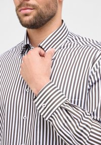 Man die de kraag van een witte shirt met verticale zwarte en grijze strepen aanpast, met een verzorgd getrimde baard en een deel van zijn gezicht in beeld.
