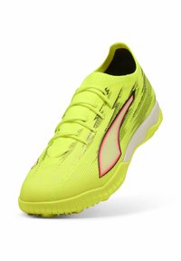 Lysegul atletiksko med en tekstureret overdel, elastiske snørebånd og en buet hvid sål, med sorte og røde detaljer.