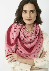 Roze sjaal met een geometrisch patroon en rode accenten, met franjes, gedrapeerd over de schouders van een persoon die een lichte blouse draagt.