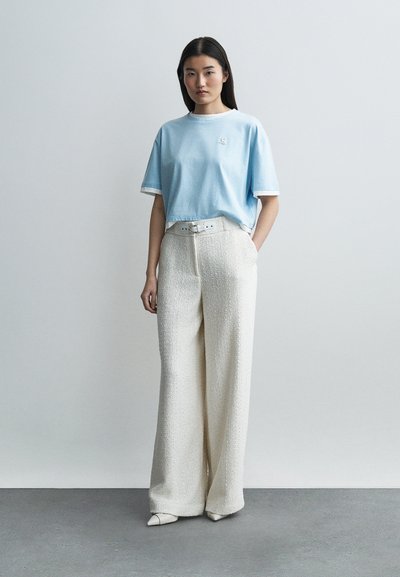 Γυναίκα που στέκεται μπροστά σε απλή λευκή επιφάνεια, φορώντας ανοιχτό μπλε κοντό τοπ, άσπρο υφασμάτινο παντελόνι με φαρδιά πόδια και ψηλή μέση, και μυτερές άσπρες γόβες.