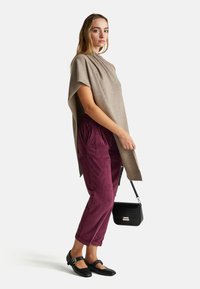 Woman wearing a beige asymmetrical poncho, burgundy corduroy pants, black buckle shoes, and holding a small black handbag.