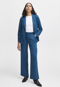 Modrý blazer s odpovídajícími širokými kalhotami, vyrobený z hladkého materiálu. Obsahuje klopový límec, otevřené kapsy a bílé tričko pod tím.