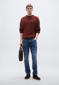 Piros bordázott pulóver, kék farmernadrág, fekete sportcipő barna talppal, barna kézitáskát tartva, semleges háttér előtt állva.