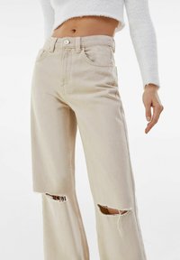 Beige wide-leg jeans with frayed knee cutouts, designed with a high waist, five pockets, and metal button closure. Soft denim texture.