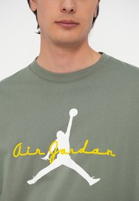 Ελιές πράσινη βαμβακερή μπλούζα T-shirt με λευκό λογότυπο Jumpman και κίτρινο κείμενο "Air Jordan" εκτυπωμένο μπροστά. Λείος, άνετος κανονισμός.