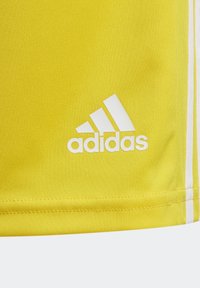 Gelbes Material mit weißem Adidas-Logo und weißen Streifen an der Seite, das einen Teil der Textur und der Nähte eines Kleidungsstücks zeigt.