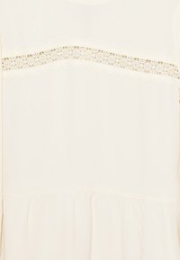 Krémový top s krajkovým pruhem přes hruď. Tkanina je hladká, se na spodní části s nařasenou strukturou.