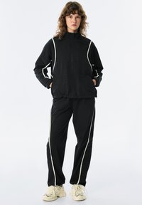 Tuta nera con strisce bianche ai lati. La giacca ha un colletto alto e una chiusura con zip. I pantaloni presentano una vestibilità comoda e tasche.