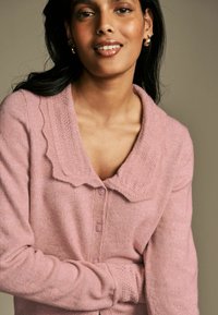 Šviesiai rožinė kardiganas su tekstūruotu apykakle, mygtukų priekyje ir ribotos rankovėmis, pagamintas iš minkštos medžiagos. Turi apvalų iškirpimą ir subtilų raštą.