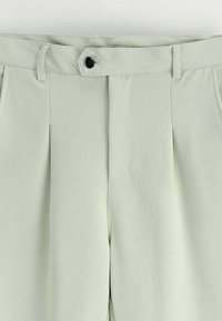 Pantalons vert clair avec une texture lisse, ornés de plis, d'une fermeture à bouton et de passants de ceinture. Design ajusté, sans poches visibles.
