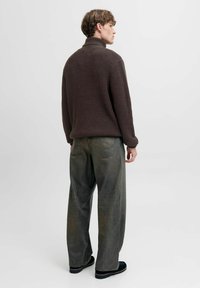 Pull en tricot marron avec col haut, associé à un jean gris-vert ample et des chaussures noires. Modèle vu de dos.