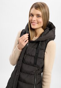 Schwarze gesteppte Weste mit Kapuze, ausgestattet mit einem Reißverschluss vorn, Seitentaschen und einem weichen beigen Pullover darunter. Texturiertes Material und schlankes Design.