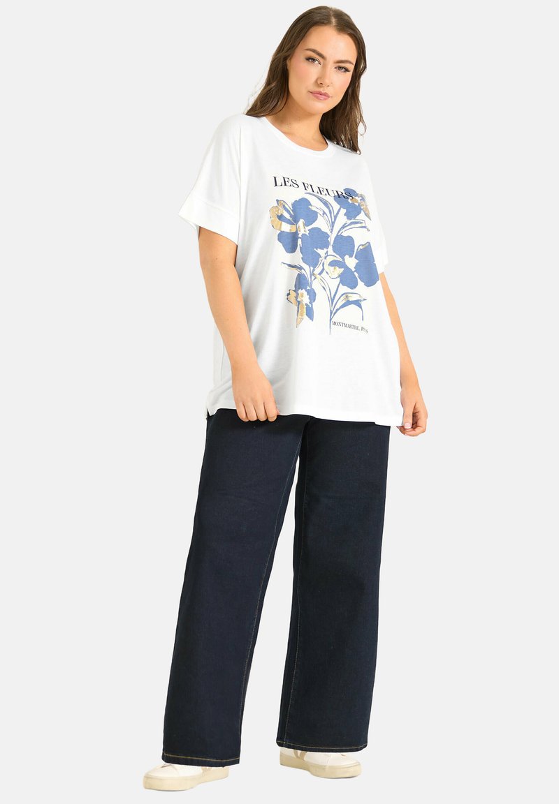 Frau trägt ein weißes oversized T-Shirt mit blauem Blumenmuster und Schriftzug, kombiniert mit weit geschnittenen dunklen Jeans und weißen Sneakers, steht vor weißem Hintergrund.