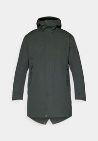 Mørkegrøn vandtæt jakke med høj krave, justerbar hætte, sidelommer og en let længere bagkant. Glat struktur og minimalistisk design.