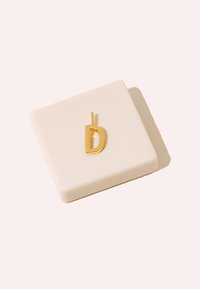 Gul bokstav "D" hänge på en beige fyrkantig bas. Hänget har en minimalistisk design med en slät yta och en framträdande form.