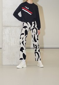Personne portant un sweat-shirt noir à manches longues FILA, un pantalon à motif abstrait noir et blanc, des chaussettes blanches et des baskets blanches épaisses, debout à l'intérieur.