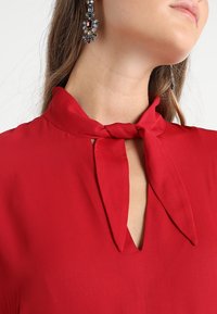 Rote Bluse mit Bindekragen, die über einen tiefen V-Ausschnitt und einen fließenden Stoff verfügt. Sie hat eine strukturierte Oberfläche und ein glattes, weiches Material.