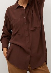 Chemise marron à boutons, avec des manches longues, deux poches sur la poitrine et une coupe décontractée. Tissu lisse avec une texture légèrement brillante.