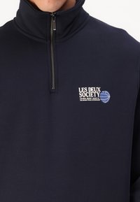 Felpa zip con collo alto di colore blu navy, con un logo bianco sulla parte superiore sinistra. Realizzata in tessuto morbido e testurizzato per garantire comfort.