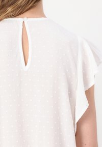 Blouse blanche texturée avec un motif délicat de points en relief, manches volantées et fermeture dos goutte d'eau sécurisée par un petit bouton.