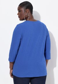 Top azul real con un patrón geométrico texturizado, escote redondo y mangas de tres cuartos. Combinado con jeans oscuros.