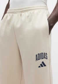 Ανοιχτό μπεζ φόρμα με ελαστική μέση, με σκούρο μπλε λογότυπο Adidas και τρεις ρίγες στη αριστερή πλευρά.