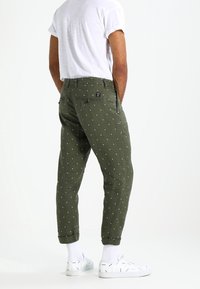 Bavlněné kalhoty olivově zelené barvy s bílým puntíkovým vzorem, zúženými nohavicemi, ohrnutými manžetami a detailem zadního kapsy, doplněné bílými teniskami.