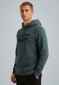 Kapuć sweatshirt u tamnozelenoj boji s prednjim kengur-džepom, s natpisom "PME LEGEND AMERICAN CLASSIC" i grafikom krila u crnoj boji.