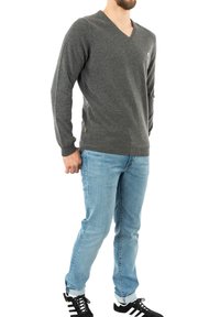 Grauer V-Ausschnitt-Pullover mit langen Ärmeln, aus weichem Stoff, kombiniert mit hellblauen Jeans und schwarzen Sportschuhen. Schlichtes Design, ohne Muster.
