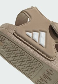 Sandale beige Adidas avec bride réglable à scratch, bandes de logo blanches, semelle texturée, et semelle intermédiaire rembourrée pour le confort.