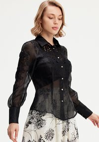 Femme portant un chemisier noir transparent avec des broderies florales et des manches bouffantes, associé à une jupe blanche ornée de motifs floraux noirs.