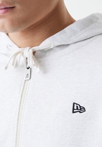 Lys grå hoodie med frontlynlås, hvide snore og et lille sort broderet logo på brystet. Blød tekstur og afslappet design.