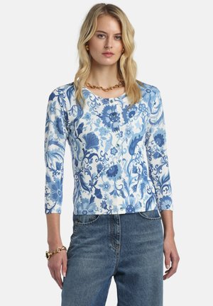 Kvinde iført en blå og hvid cardigan med blomsterprint og knapper, trekvartærmer, kombineret med højtaljede blå jeans og guldsmykker.