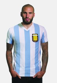 Camisa de manga corta con escote en V, que presenta rayas verticales alternantes en azul claro y blanco, con un emblema bordado en el lado izquierdo del pecho.