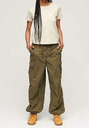 Persoon draagt een wit shirt met korte mouwen, olijfgroene cargobroek met zakken en beige werkschoenen, staat met handen in de zakken.