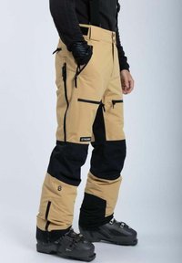 Beige og sorte skibukser med flere lynlåslommer, justerbare stropper og et vandafvisende stof, parret med sorte skistøvler.