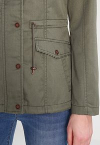 Zbliżenie na oliwkowo-zieloną kurtkę z przednimi zatrzaskami, kieszenią z patką oraz regulowanym ściągaczem, noszoną na niebieskich dżinsach z widoczną dłonią.