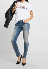 Camiseta blanca con logo "REPLAY" y rayas de colores, combinada con jeans ajustados de color azul claro y tacones altos negros. Chaqueta de cuero colgada en el brazo.