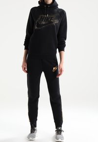 Svart Nike-hoodie med en guldglittrig logotyp, i kombination med svarta sweatpants som har en guldlogotyda. Mjuk material, avslappnad passform.
