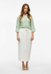 Camicia verde chiaro con maniche a tre quarti, abbinata a pantaloni bianchi a vita alta e a gamba larga. Sandali semplici da infilare completano l'outfit.