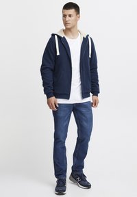 Granatowa bluza z kapturem na zamek z kremowym wykończeniem, elastycznymi mankietami i dwiema przednimi kieszeniami, w połączeniu z niebieskimi dopasowanymi dżinsami i czarnymi butami biegowymi.