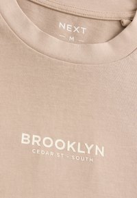 Béžové bavlnené tričko s okrúhlym výstrihom, na prednej strane s bielym potlačeným textom "BROOKLYN CEDAR ST - SOUTH". Hladká textúra, neformálny dizajn.