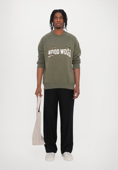 Olivovo zelená mikina so nápisom "WOOD WOOD", raglánové rukávy; nosená s čiernymi nohavicami a svetlými teniskami, drží béžovú tašku.