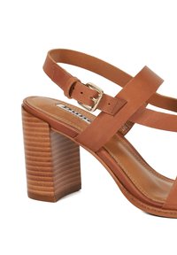 Brown leather heeled sandal with a cross strap design, adjustable buckle, and a ribbed wooden heel. Smooth texture and rounded toe.